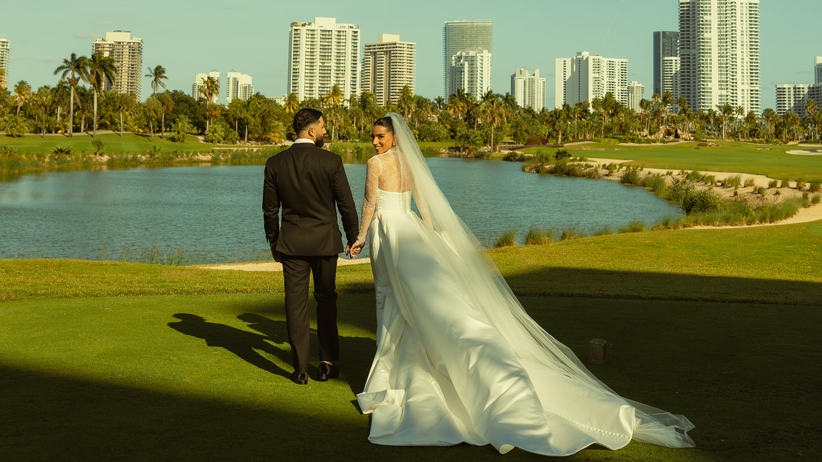 Smiling Bride holding hands with Groom Portraits on Golf Course Aventura Florida with Miami Skyline in the background. Documentary Wedding Photographer.