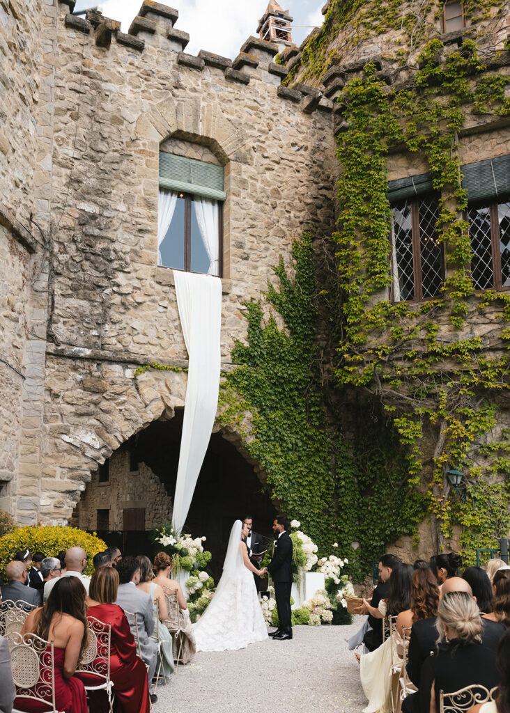 A bride and groom exchanging vows at an outdoor altar adorned with lush white florals in front of an ivy-covered stone castle in Spain, demonstrating how hiring a wedding planner ensures flawless execution for historic destination venues.