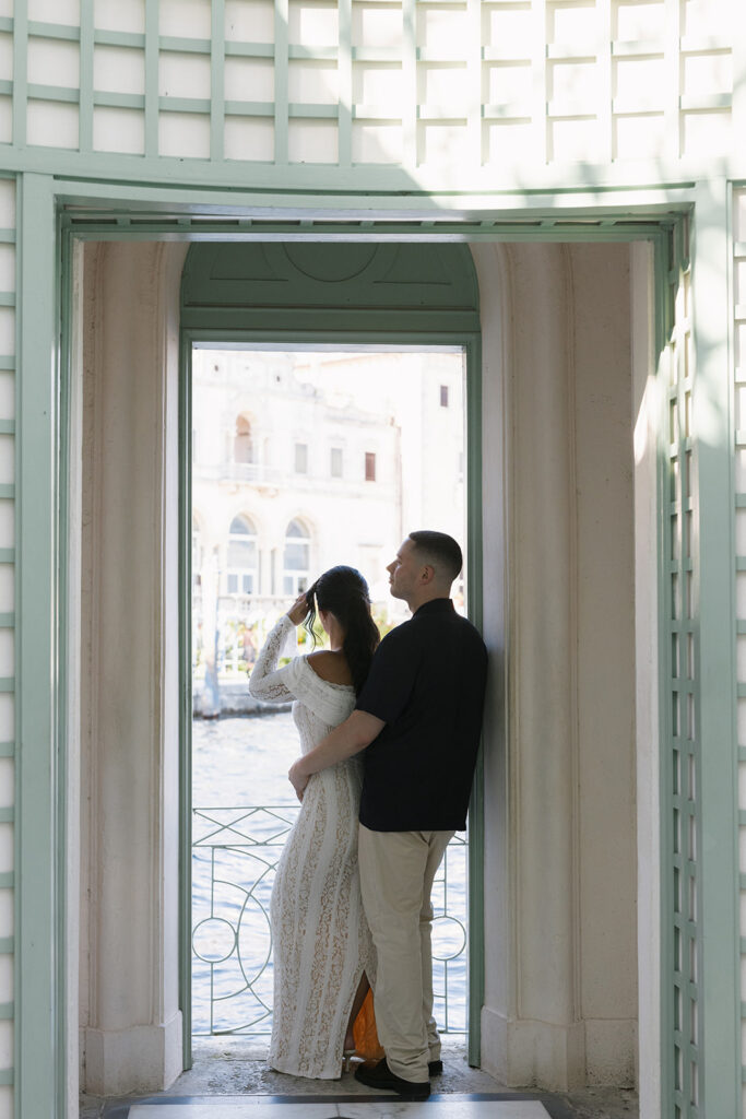 An engaged couple embracing in an elegant, green-latticed archway overlooking the water, showing how hiring a wedding planner helps couples discover and secure exclusive destination venues.